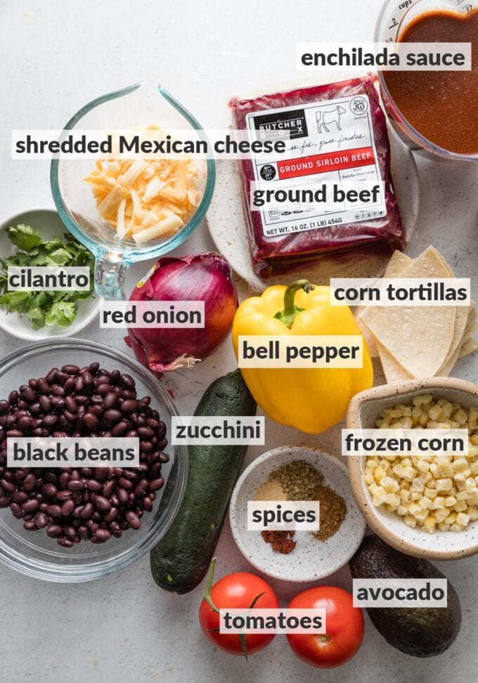 Labeled overhead view of ground beef, enchilada sauce, a yellow bell pepper, a zucchini, red onion, cheese, black beans, and spices, ready to cook into an enchilada skillet.