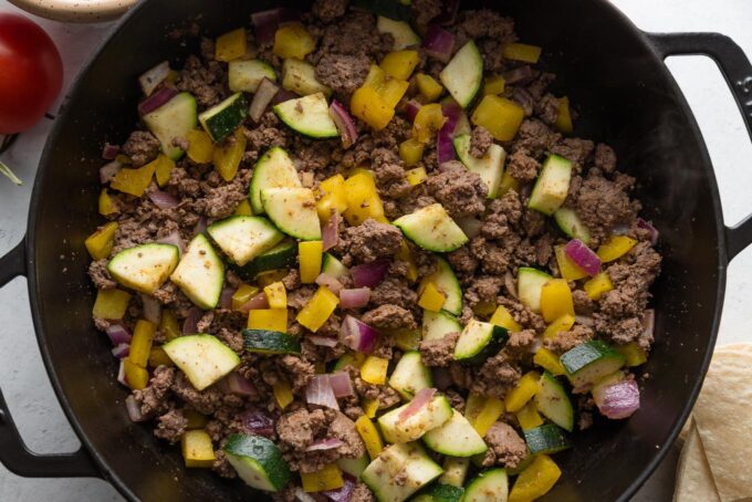 Ground beef browned in a skillet with bell pepper, zucchini, and red onion.