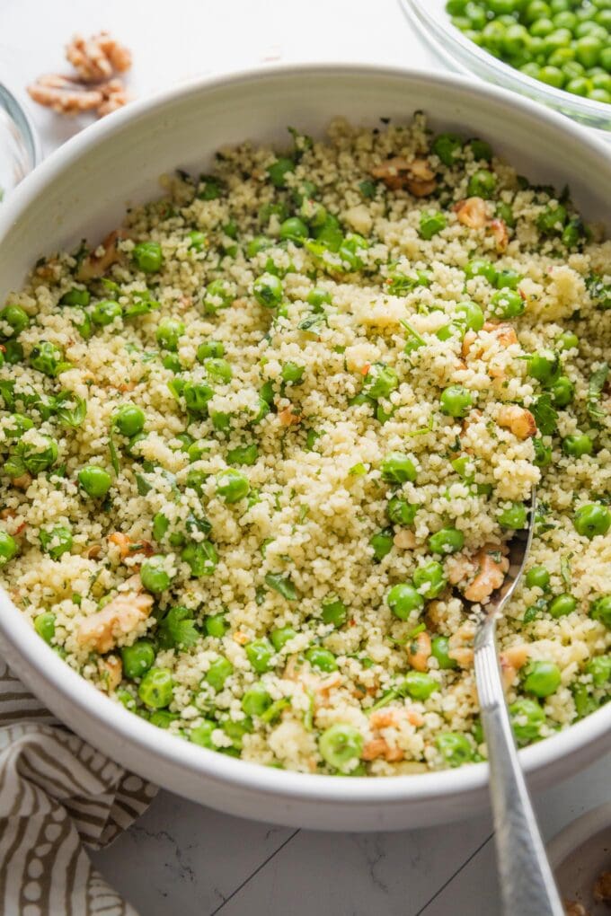 Angled view of a serving spoon lifting a portion of couscous salad from a white serving bowl.