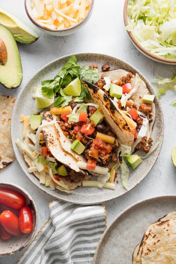 Countertop set with a plate of ground beef and black bean tacos, extra cheese, lettuce, avocado, and tortillas.