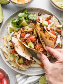 A woman's hand picking up a taco filled with a ground beef and black bean mixture, lettuce, tomato, and avocado.