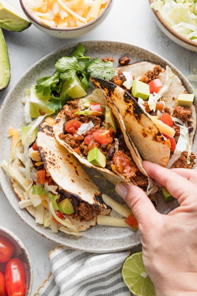 A woman's hand picking up a taco filled with a ground beef and black bean mixture, lettuce, tomato, and avocado.