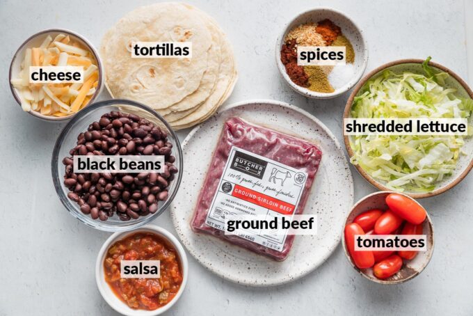 Labeled overhead photo of ground beef, drained and rinsed black beans, salsa, spices, tortillas, tomato, lettuce, and shredded Mexican cheese.