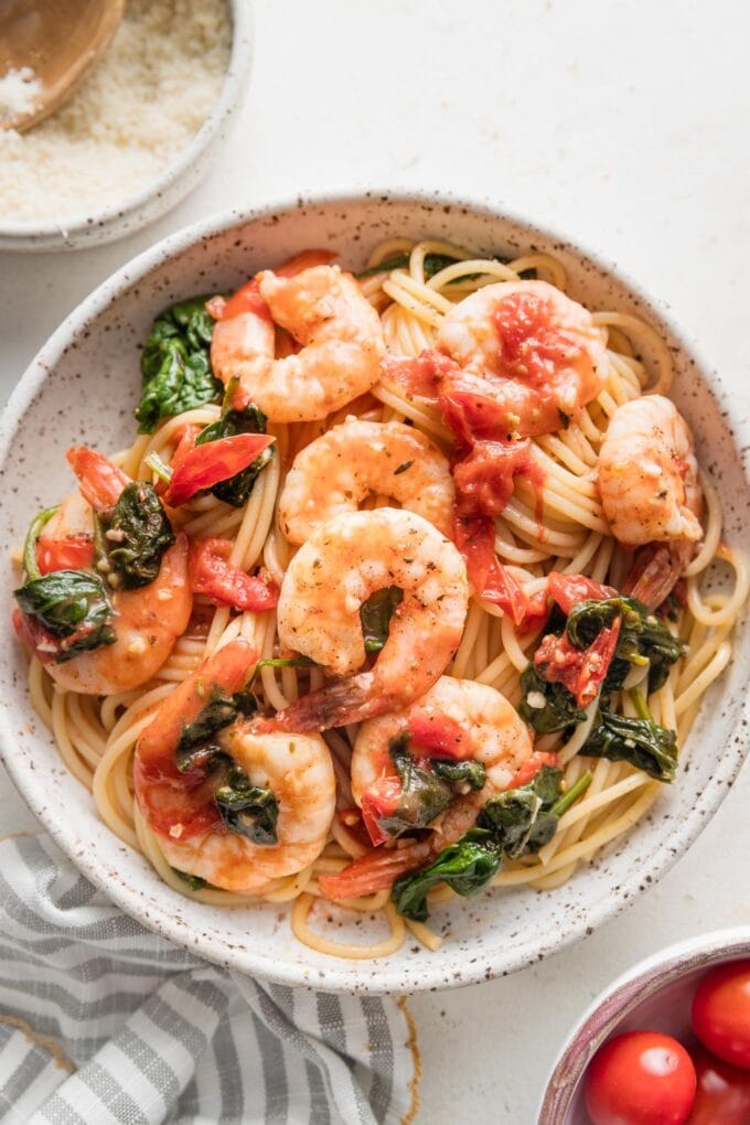 Bowl of tomato spinach shrimp pasta with a striped napkin and extra cherry tomatoes on the counter nearby.