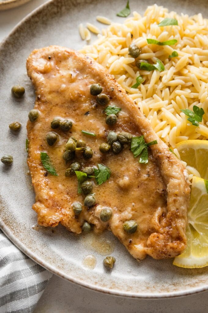 Plate of lemon piccata chicken served with extra capers, parsley, and orzo pasta.