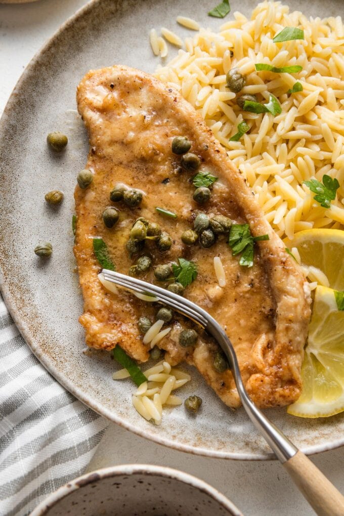 Close up of a fork slicing off a piece of tender chicken piccata, with a side of orzo.