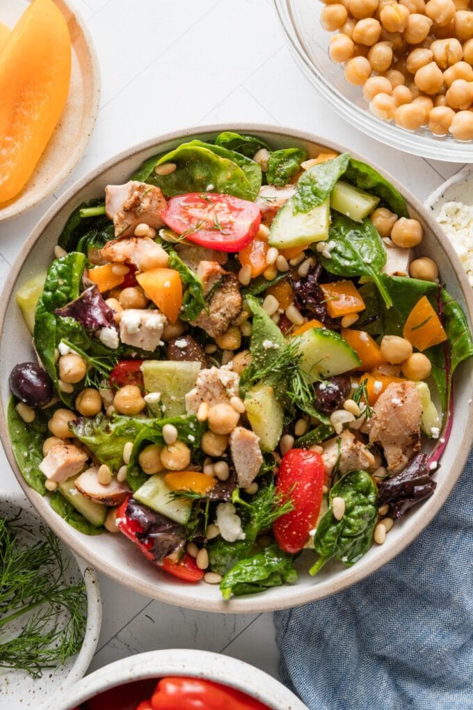 Countertop set with a lunch portion of Mediterranean chopped salad, with bowls full of extra chickpeas, orange bell pepper, fresh dill, and cherry tomatoes nearby.
