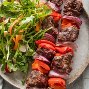 Plate with juicy steak kabobs with bell pepper and onion served with an arugula carrot salad.
