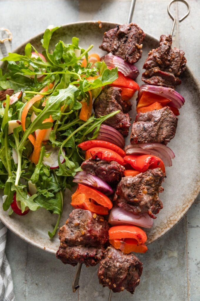 Plate with juicy steak kabobs with bell pepper and onion served with an arugula carrot salad.