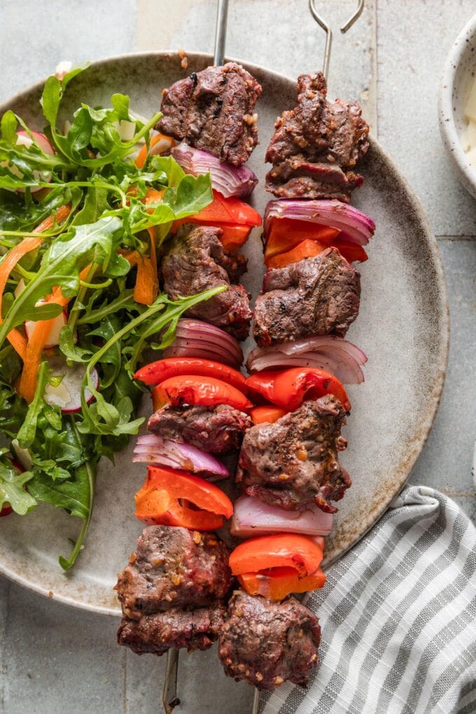 Close up of steak kabobs on a plate with an arugula radish and carrot salad.