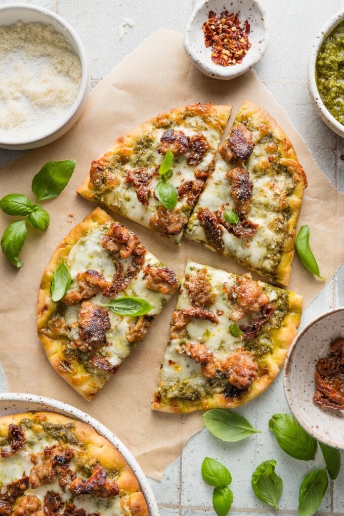 A naan flatbread pizza with pesto, sausage, sun-dried tomatoes, and cheese, sliced into four pieces and ready to serve.