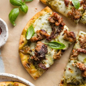 Close up of a slice of pesto sausage flatbread with fresh basil leaves on top as a garnish.