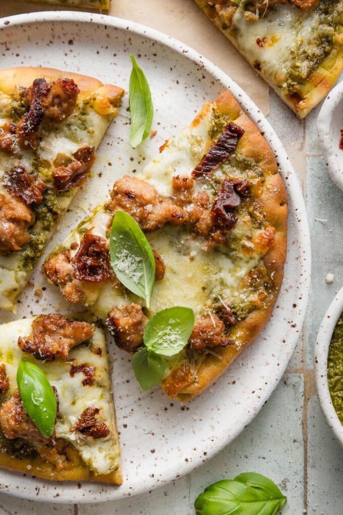 Small white ceramic plate holding slices of pesto sausage flatbread with red pepper flakes and fresh basil for garnish.
