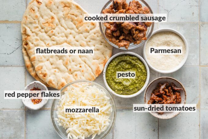 Labeled overhead photo of plain naan, pesto, mozzarella and Parmesan cheese, sun-dried tomatoes, red pepper flakes, and cooked crumbled Italian sausage.
