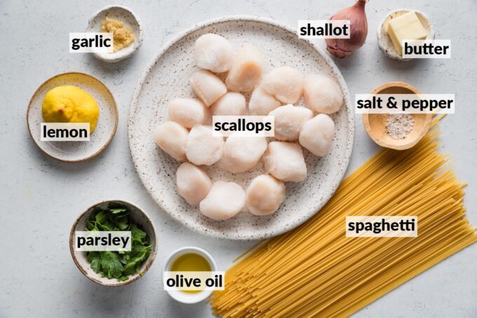 Labeled overhead photo of sea scallops, uncooked spaghetti, a shallot, garlic, butter, olive oil, a lemon, and kosher salt and black pepper.