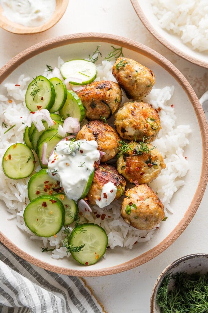 Bowl of chicken zucchini meatballs served over rice with a cucumber shallot salad and Tzatziki sauce.