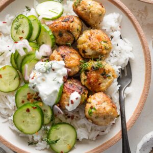 Bowl full of chicken zucchini meatballs served over rice with a cucumber shallot salad, fresh dill, and Tzatziki sauce.