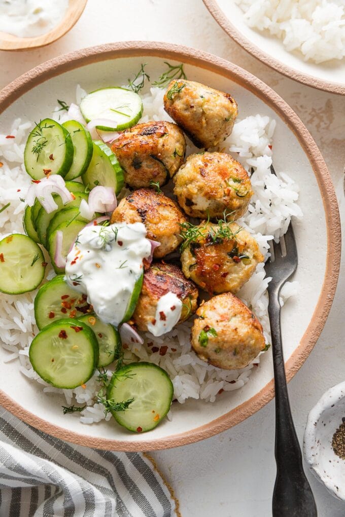 Bowl full of chicken zucchini meatballs served over rice with a cucumber shallot salad, fresh dill, and Tzatziki sauce.