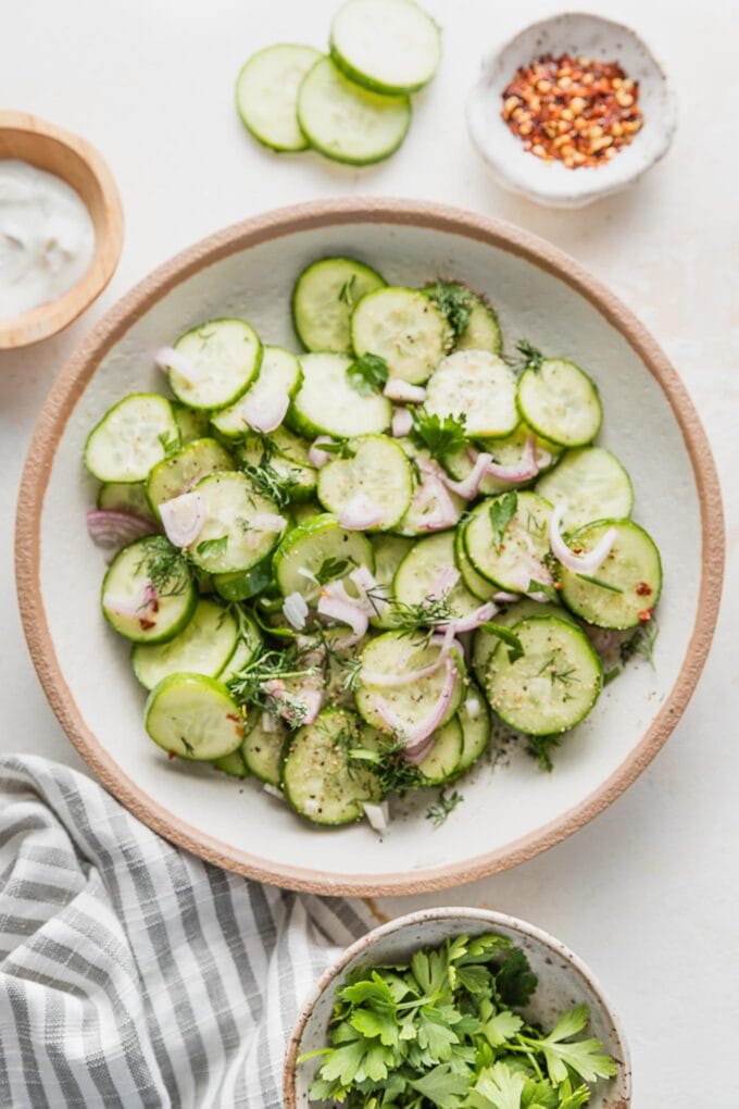 Countertop set with a bowl of cucumber shallot salad with extra fresh herbs, Tzatziki sauce, and cucumber slices scattered nearby.