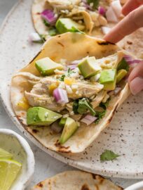 Hand holding a salsa verde chicken taco with avocado and diced onion, over a small ceramic plate.