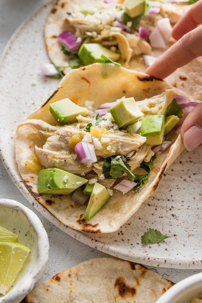Hand holding a salsa verde chicken taco with avocado and diced onion, over a small ceramic plate.