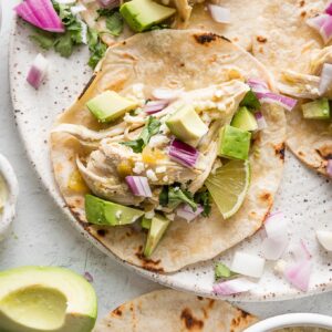 Plate full of slow cooker salsa verde chicken tacos with avocado, lime wedges, cilantro, and Cotija.