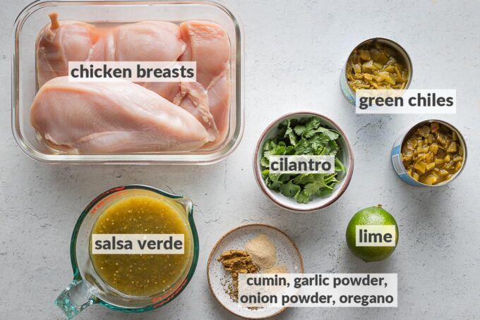 Labeled photo of ingredients for Crockpot salsa verde chicken: chicken breasts, salsa verde, two cans mild green chiles, lime, cilantro, and spices including cumin, Mexican oregano, garlic powder, and onion powder.