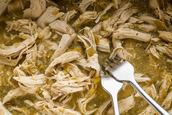 Two forks shredding chicken breasts slow cooked in salsa verde with chiles and spices.