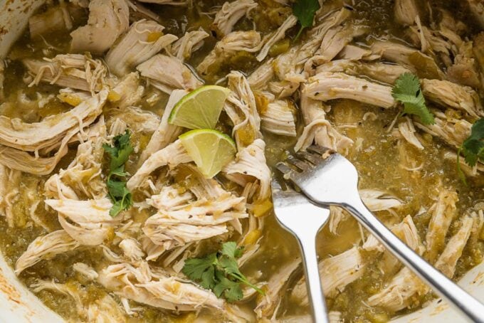 Large slow cooker bowl full of salsa verde chicken with lime wedges and cilantro leaves for added flavor.