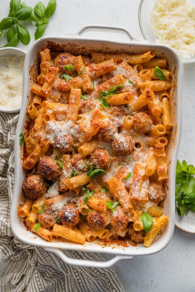 Countertop set with a large casserole dish full of baked rigatoni and meatballs garnished with fresh basil leaves, with extra grated Parmesan and parsley scattered nearby.