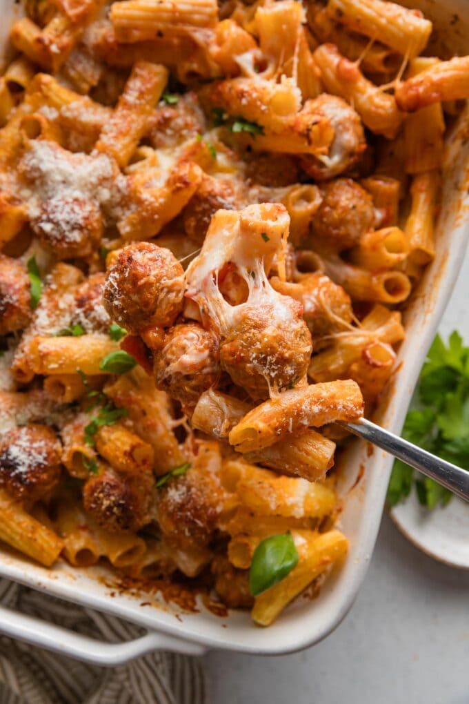 Close up of a spoon serving a cheesy meatball out of a baked pasta dish.