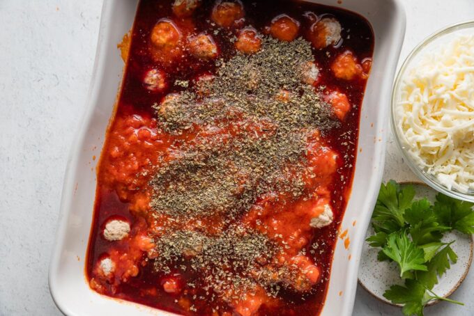 Casserole dish filled with marinara sauce, frozen meatballs, a smidge of red wine, and seasonings sprinkled on top.