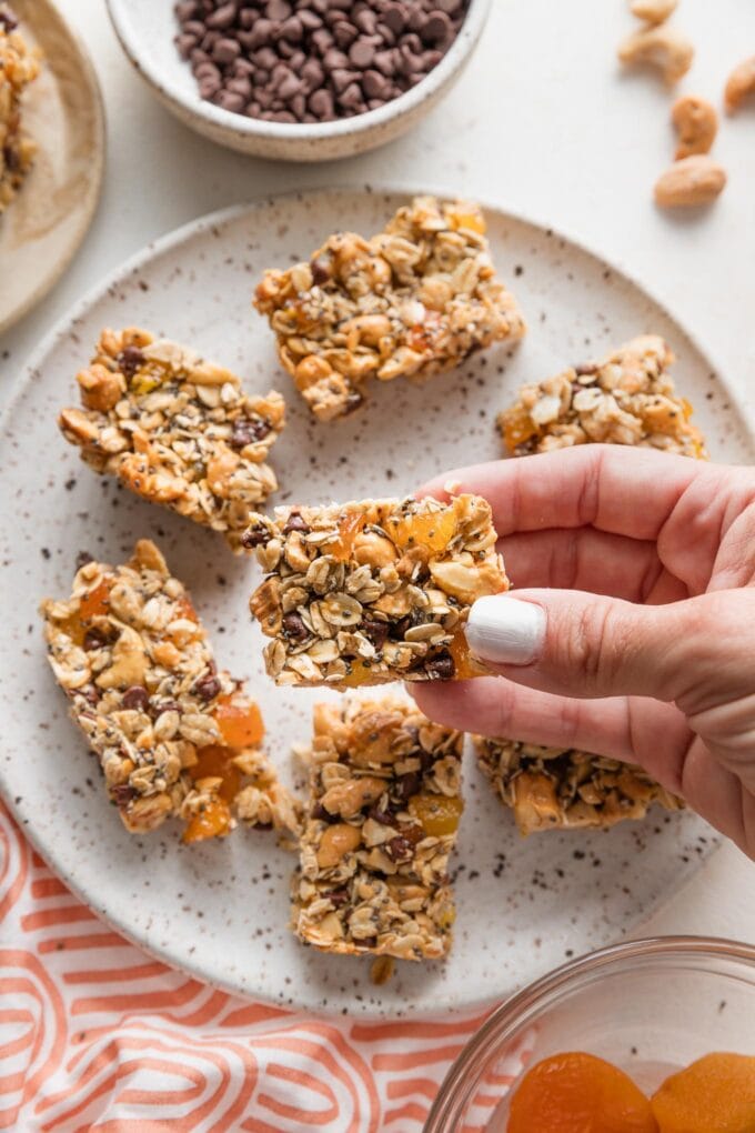 Close up of a hand holding a chocolate cashew apricot snack bar over a plate filled with them.