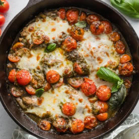 Cast iron skillet full of pesto baked chicken breasts with cherry tomatoes, shallots, garlic, and melted mozzarella.