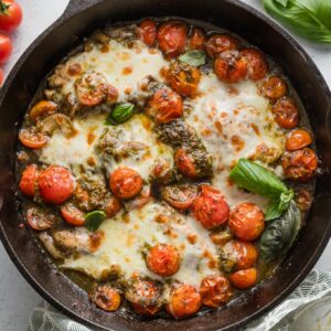Cast iron skillet full of pesto baked chicken breasts with cherry tomatoes, shallots, garlic, and melted mozzarella.