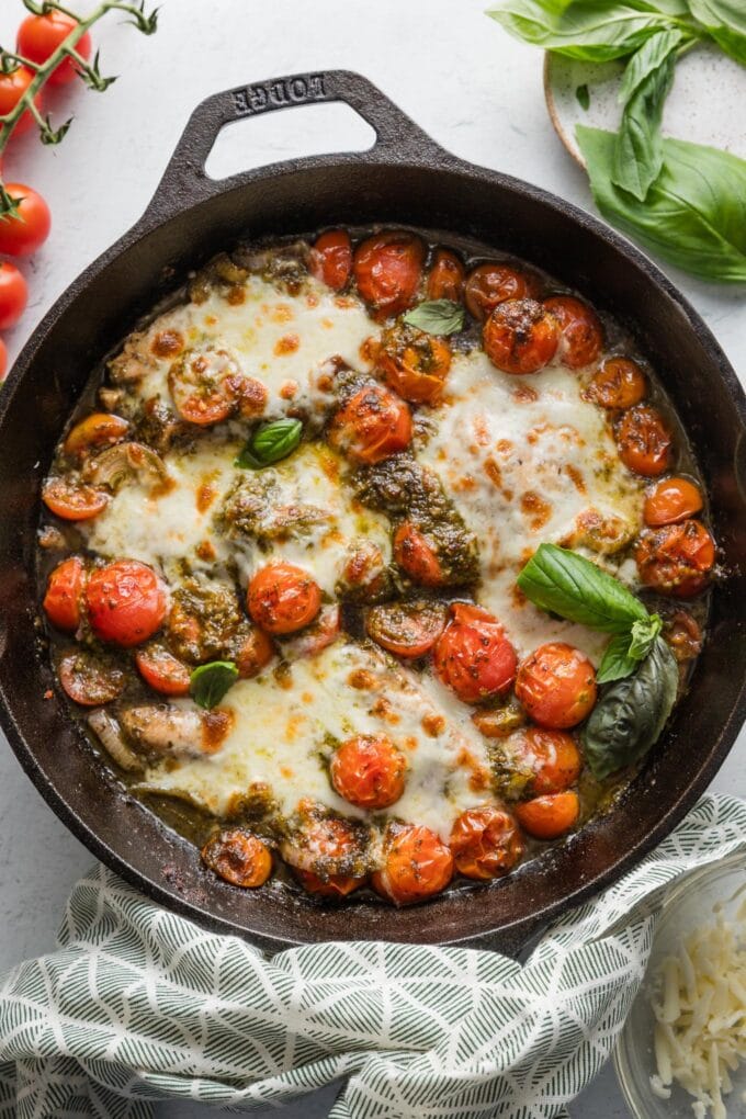 Cast iron skillet full of pesto baked chicken breasts with cherry tomatoes, shallots, garlic, and melted mozzarella.