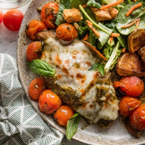 Plate with pesto baked mozzarella chicken, burst cherry tomatoes, roasted red potatoes, and a simple salad.