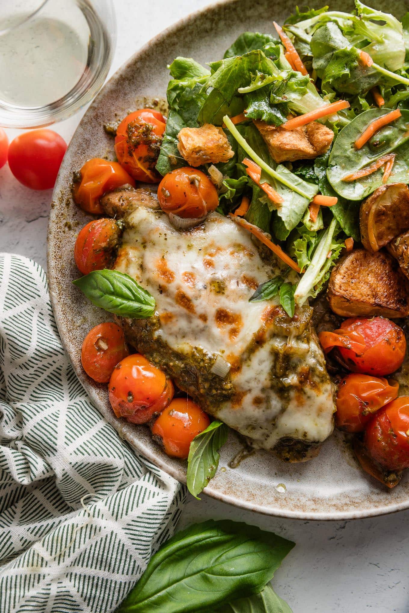 Plate with pesto baked mozzarella chicken, burst cherry tomatoes, roasted red potatoes, and a simple salad.