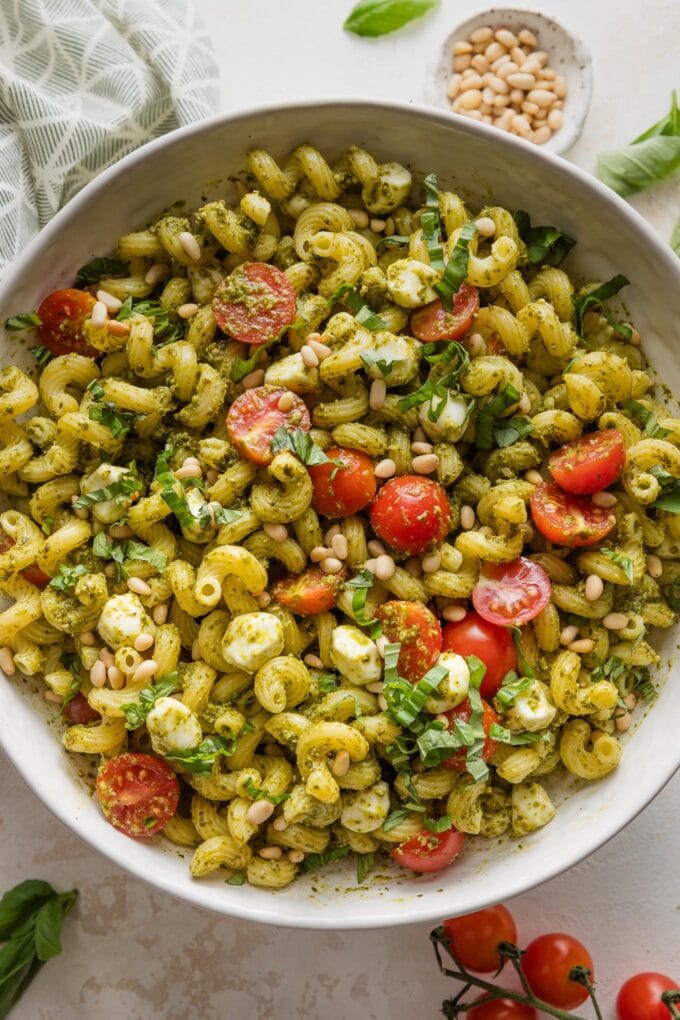 Close up of pesto pasta salad with pine nuts, mozzarella, and tomatoes in a large serving bowl.
