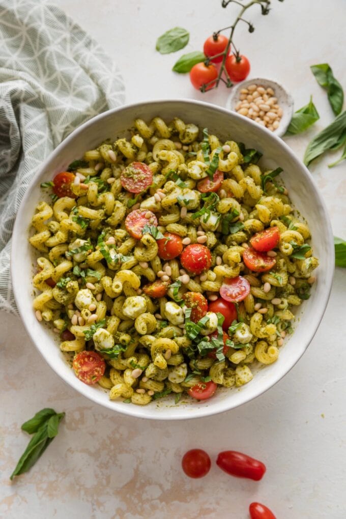 Countertop set with a large bowl of pesto pasta salad with extra basil leaves and cherry tomatoes scattered nearby.