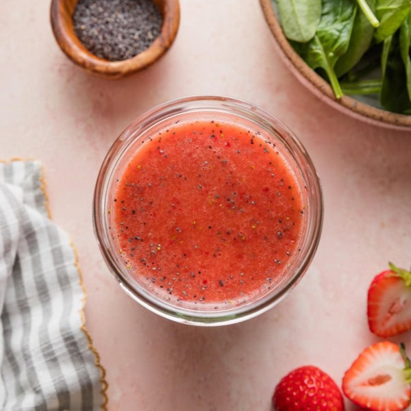 Pink countertop with a small glass jar full of homemade strawberry salad dressing with poppy seeds, with a clean striped kitchen towel, extra fresh strawberries, poppy seeds, and baby spinach leaves in bowls nearby.