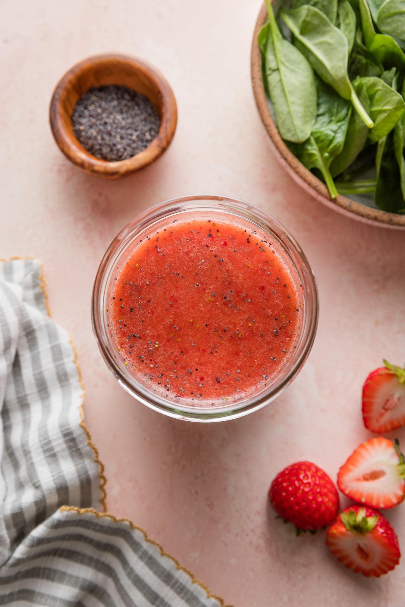 Pink countertop with a small glass jar full of homemade strawberry salad dressing with poppy seeds, with a clean striped kitchen towel, extra fresh strawberries, poppy seeds, and baby spinach leaves in bowls nearby.
