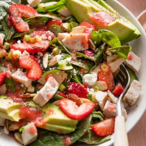 Angled view of a countertop set with a large bowl of spinach strawberry salad topped with strawberry dressing and fresh cracked black pepper.