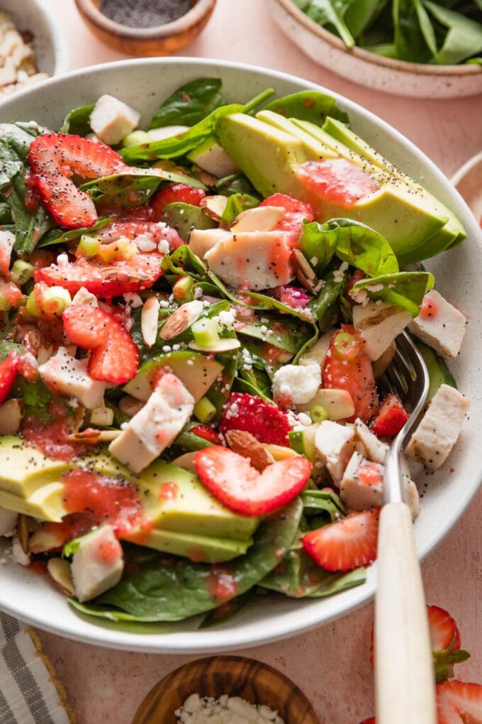 Angled view of a countertop set with a large bowl of spinach strawberry salad topped with strawberry dressing and fresh cracked black pepper.