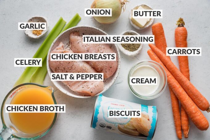 Labeled overhead image of chicken breasts seasoned with kosher salt and black pepper, whole carrots, celery, a yellow onion, fresh minced garlic, chicken broth, cream, dried Italian seasoning, and a can of store-bought biscuits.