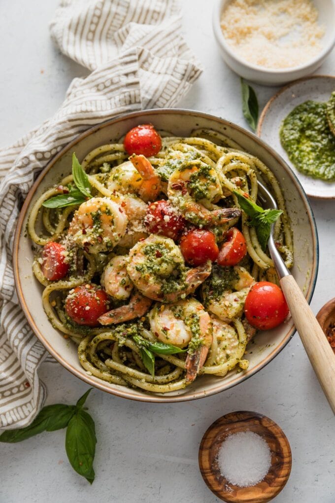 Bowl of creamy pesto shrimp served over pasta with cherry tomatoes and a lot of extra pesto and Parmesan.