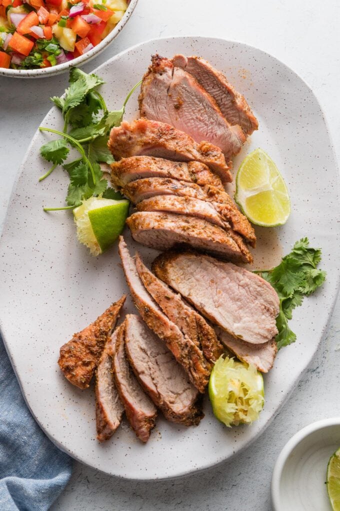 Slices of grilled pork tenderloin arranged on a platter with cilantro leaves and lime wedges.