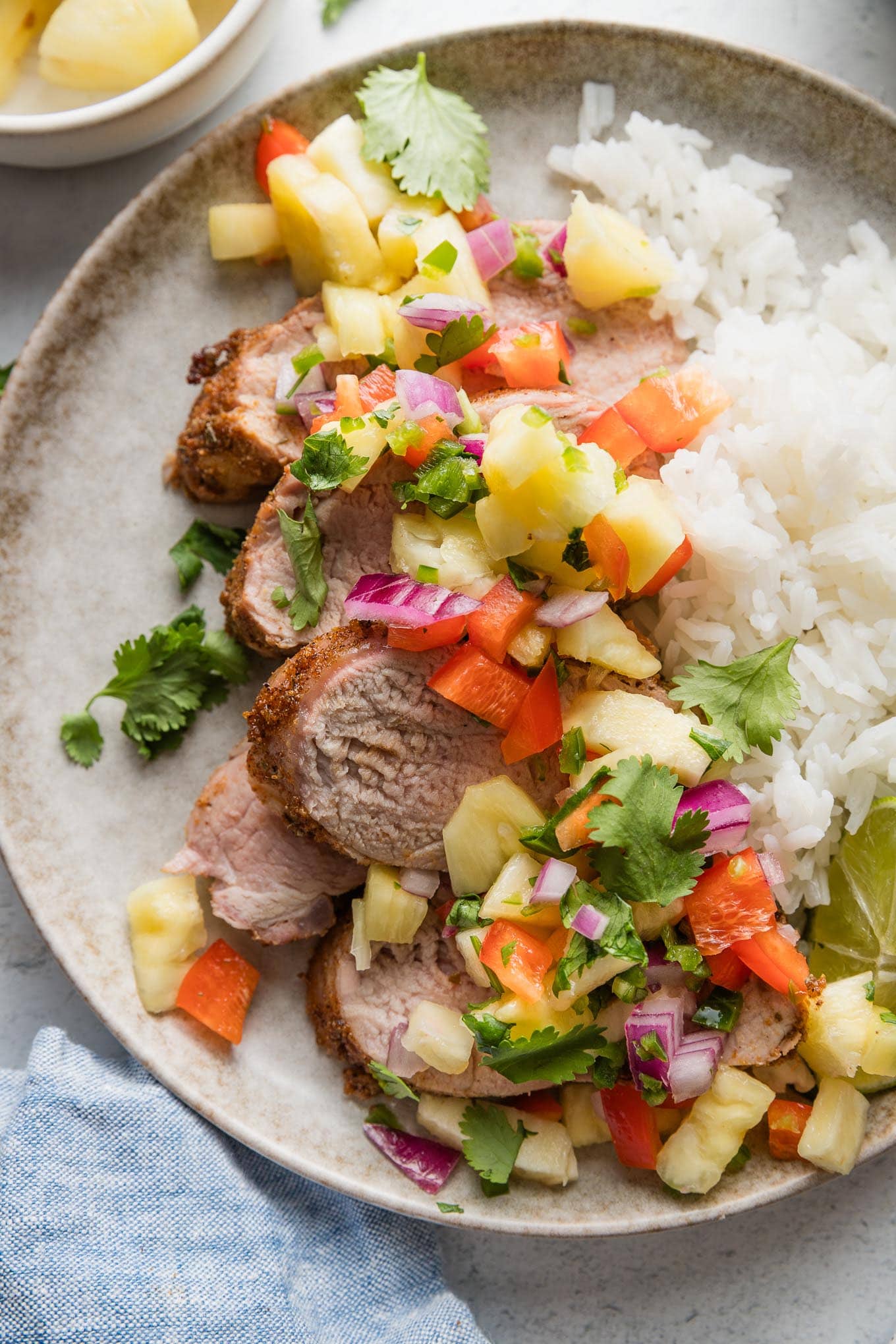 Plate with slices of grilled, seasoned pork tenderloin topped with a salsa of fresh pineapple, bell pepper, red onion, cilantro, and lime juice, served over white rice.