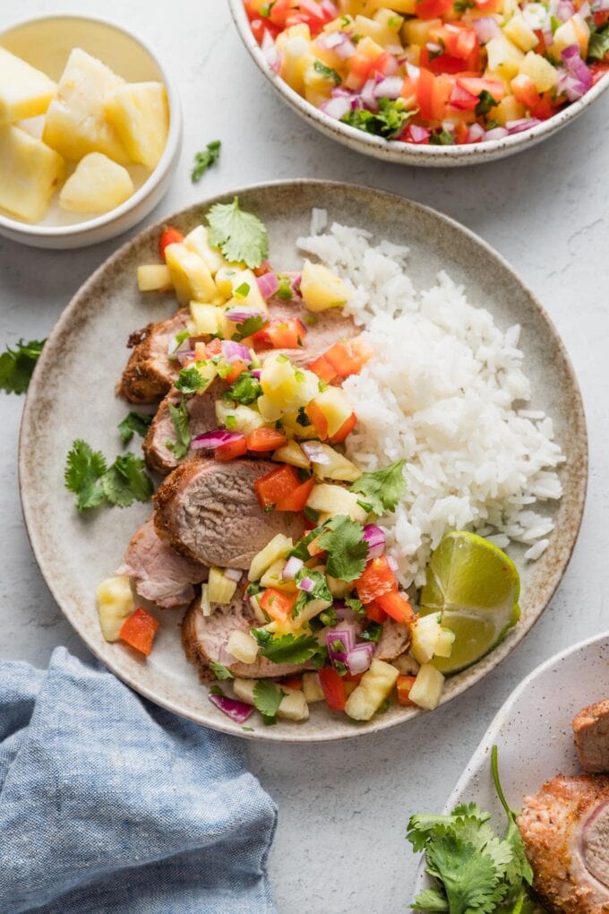 Overhead view of a small dinner plate presented with a bed of white rice, grilled pork tenderloin, and pineapple salsa, with a pretty blue kitchen cloth and extra salsa nearby on the counter.