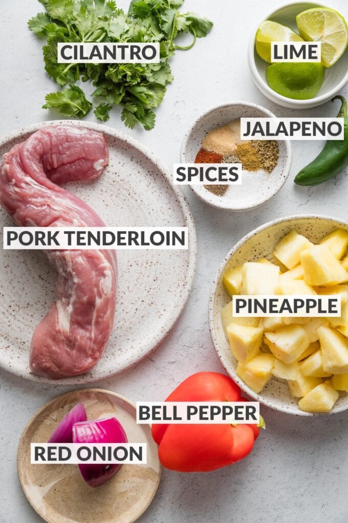 Labeled overhead image of an uncooked pork tenderloin, fresh pineapple chunks, a red bell pepper, part of a red onion, one jalapeno pepper, fresh lime and cilantro, and spices.